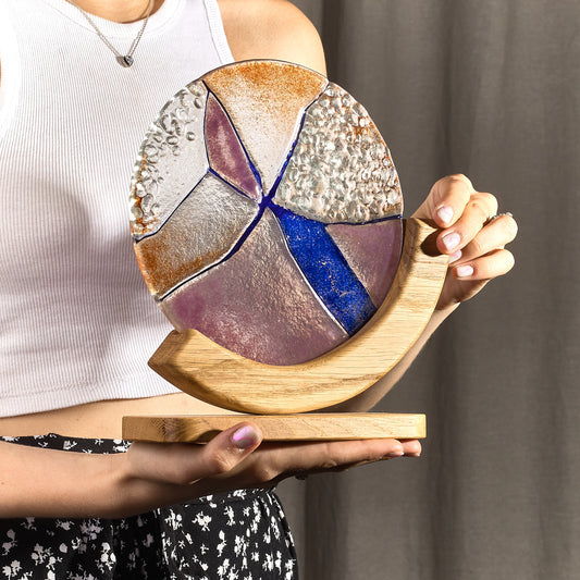 A person holding a round stained glass decor piece with a geometric pattern, featuring earthy tones and a bold streak of deep cobalt blue, mounted on a curved oak base.