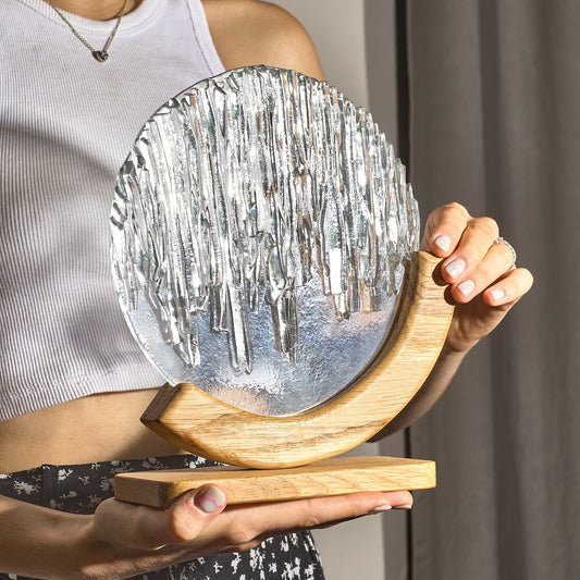 A round stained glass panel with a cascade of icicles held in a person's hand, mounted on a curved wooden base.