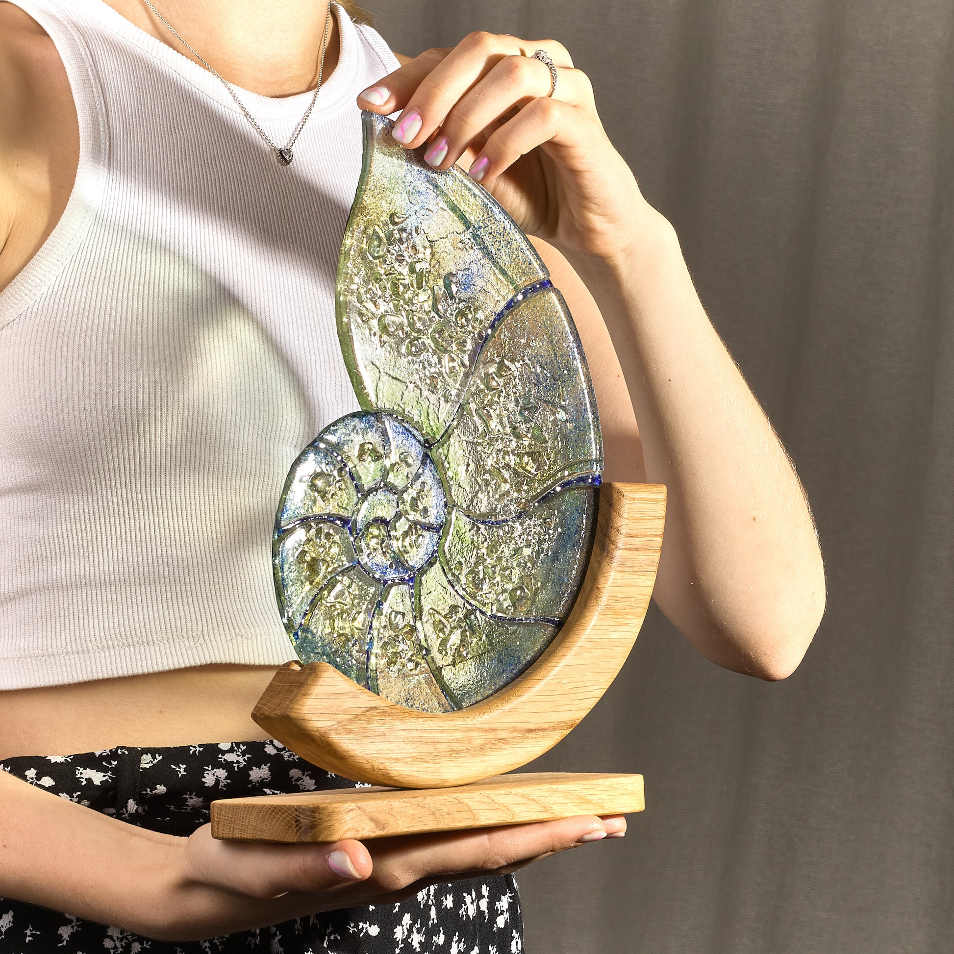 A stained glass stand in the shape of a nautilus shell with green, blue, and gold colors, held by a person. It stands on a wooden base.