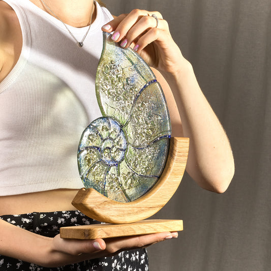 A stained glass stand in the shape of a nautilus shell with green, blue, and gold colors, held by a person. It stands on a wooden base.