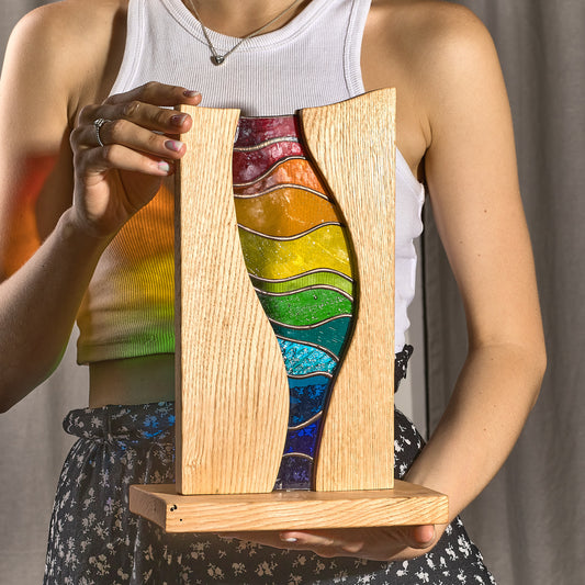 A person holding a stained glass stand with a rainbow spectrum design, featuring curved lines in red, blue, green, and yellow colors, framed by an oak wood structure.