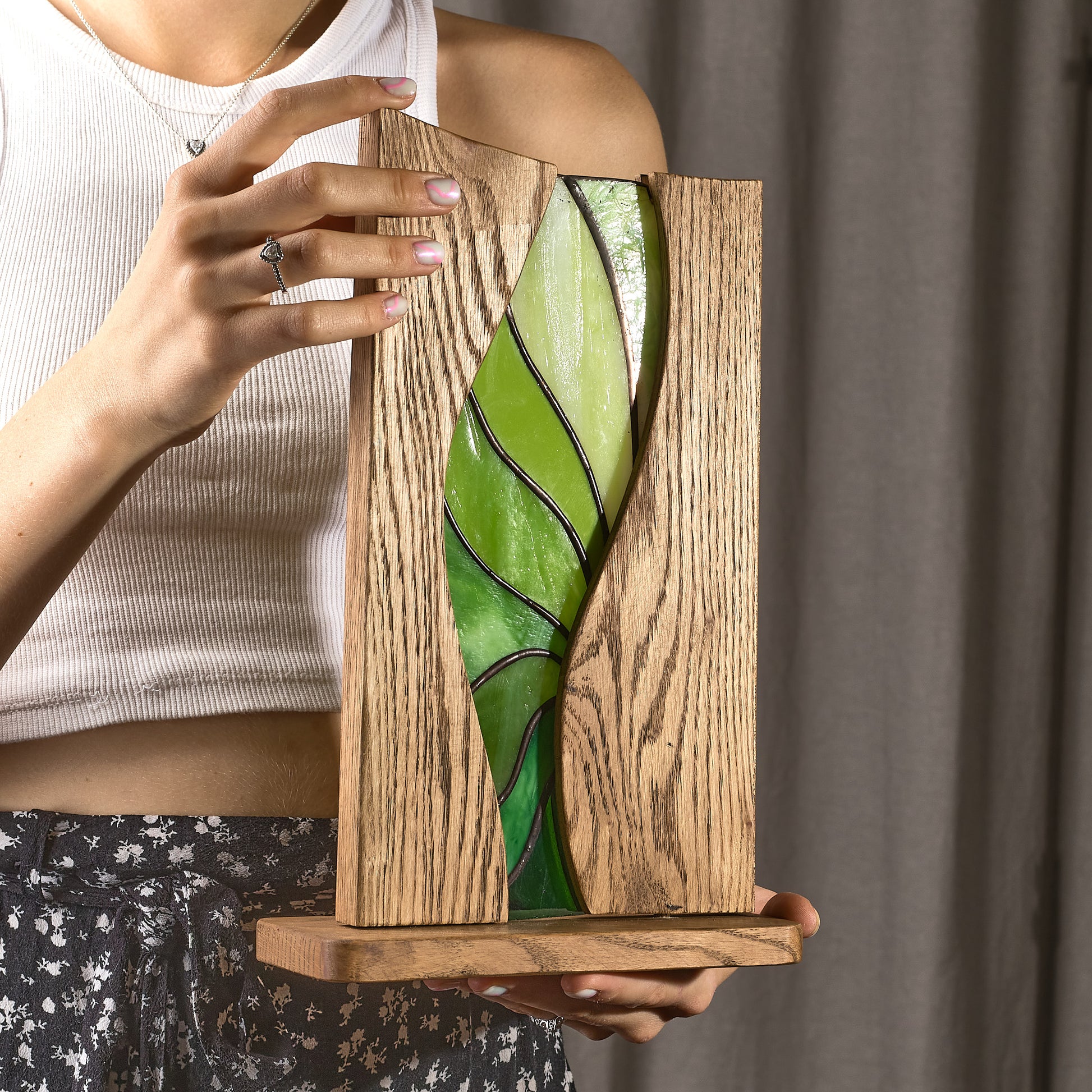 A person holding a freestanding stained glass art piece with abstract curved lines in emerald, moss, and lime hues, framed in rich oak wood.