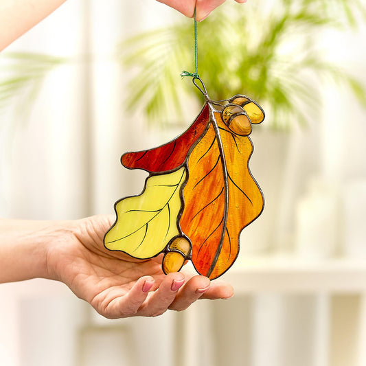 A hand holding a stained glass suncatcher featuring a trio of colorful oak leaves with acorns, with a background of a plant and furniture.