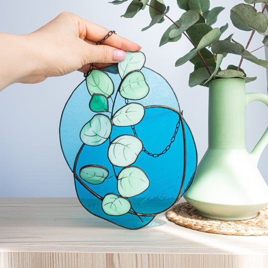 A handcrafted stained glass panel suncatcher with a vibrant eucalyptus branch design on a blue background, held by a hand against a wooden surface with a green vase and a plant in the backdrop.