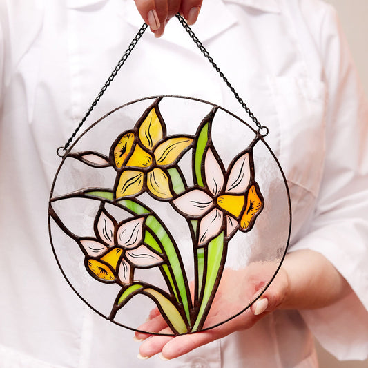 A hand holding a round stained glass panel featuring a white and yellow narcissus flower design, with a metal chain attached for hanging.