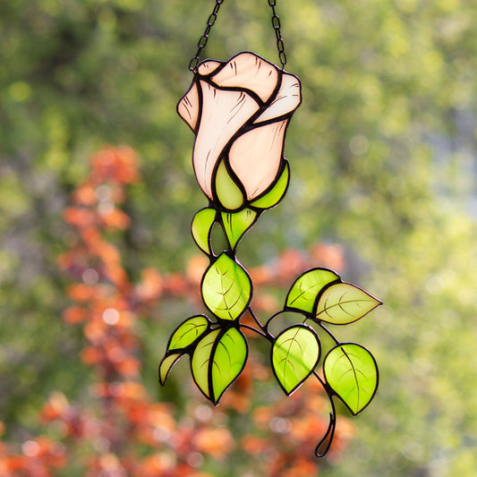 A handcrafted stained glass suncatcher in the shape of a rose flower with green leaves, hanging against a blurred background of autumn leaves.