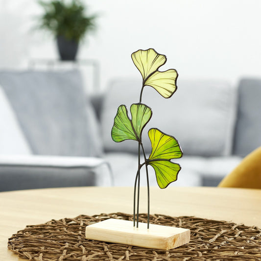 A handcrafted stained glass Ginkgo Leaf table stand decoration with a metal stand, placed on a wooden platform, set against a cozy home background.