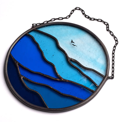 A circular stained glass suncatcher with varying shades of blue and a bird silhouette, designed to be hung by a chain.