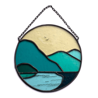 A round stained glass suncatcher featuring geometric shapes in shades of green and blue, resembling mountain ranges and a lake, with a chain for hanging.