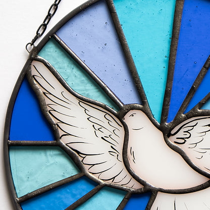 Stained glass suncatcher featuring a white dove with wings extended, set against a blue background with rays of light originating from the dove.