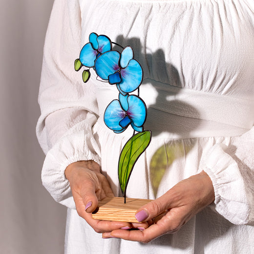A person holding a stained glass artwork of a blue orchid flower on a wooden platform.