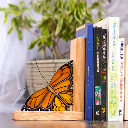 A pair of handmade stained glass bookends featuring monarch butterflies, placed on a bookshelf with books and a plant in the background.