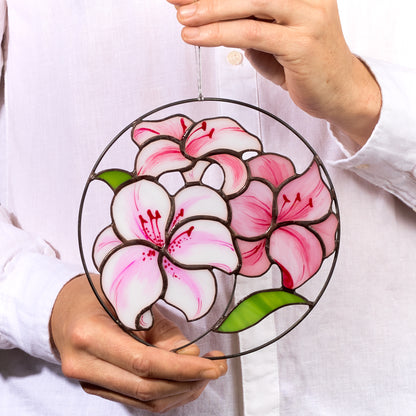 A round stained glass suncatcher featuring a floral design with pink and purple hues, held in a person's hand.