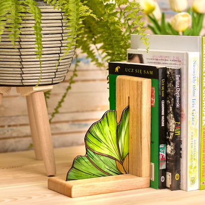 A pair of green stained glass Ginkgo leaf bookends with oak wood bases, holding up a row of books on a wooden shelf.