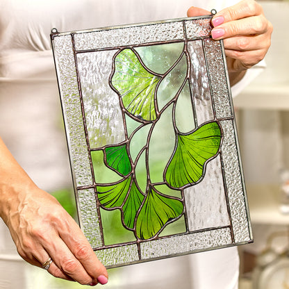 A handcrafted stained glass panel with a depiction of ginkgo leaves in vibrant green tones, held by a person.