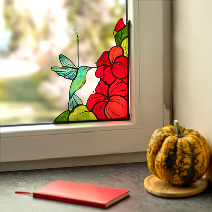 Stained glass window corner panel with a design of a hummingbird and red hibiscus flowers, placed in a window with a suction cup.