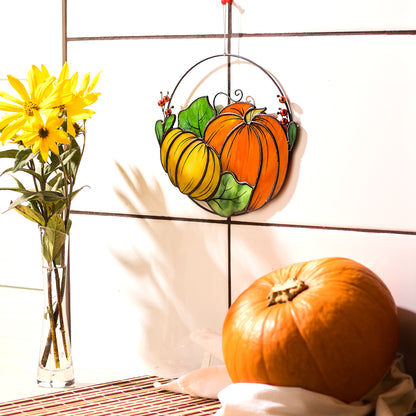 A handmade stained glass suncatcher featuring two pumpkins in shades of yellow and orange with green leaves and red berries, hanging on a wall above a pumpkin patch.