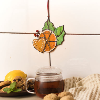 A stained glass suncatcher with a vibrant orange slice design, displayed in front of a window with light coming through, casting colorful reflections. The suncatcher is accompanied by a gingerbread heart and festive holly leaves with red berries.