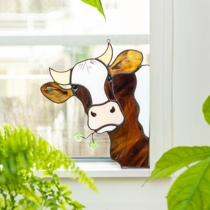A handcrafted stained glass window decoration featuring a whimsical cow design, placed in a window corner with plants in the background.