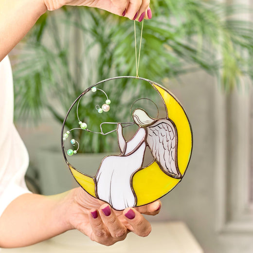 A hand holding a stained glass suncatcher featuring an angel playing a flute, with a crescent moon and green vines with pearl beads.