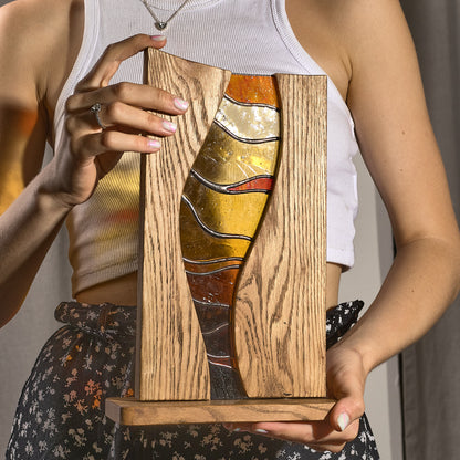 A person holding a stained glass stand with amber, orange, and gold hues, framed by oak wood.