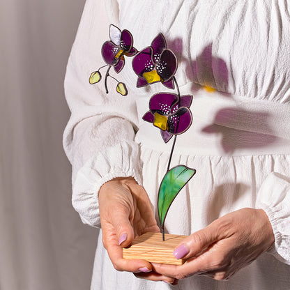 A person holding a stained glass orchid with deep purple petals and golden-yellow accents on a wooden platform.