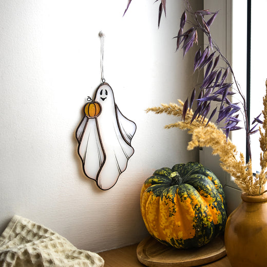 A handmade stained glass ghost with a pumpkin hanging on a wall, next to a pumpkin patch and a plant in a brown vase.