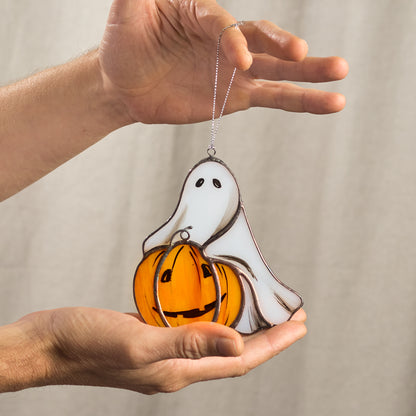 A hand holding a stained glass suncatcher featuring a white ghost with a pumpkin.