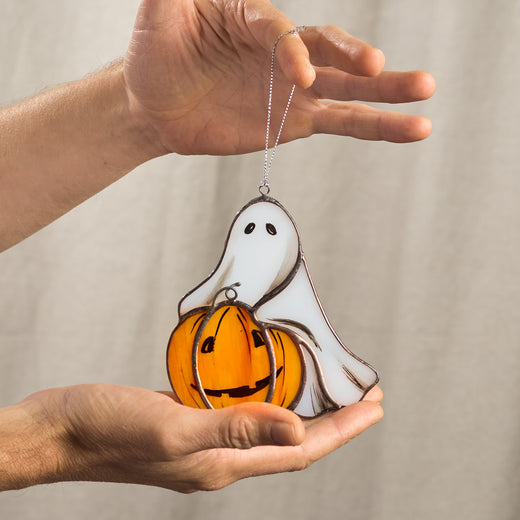 A hand holding a stained glass suncatcher featuring a white ghost with a pumpkin.