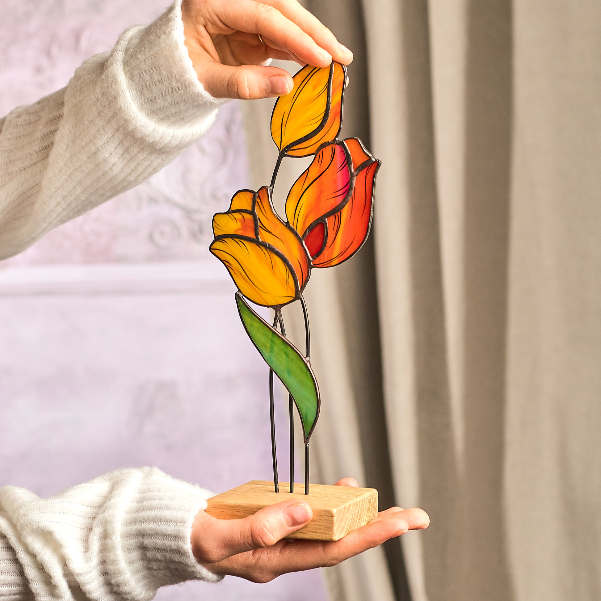 A handcrafted stained glass representation of tulips in orange and pink colors on a wooden platform, held by a person against a blurred background.