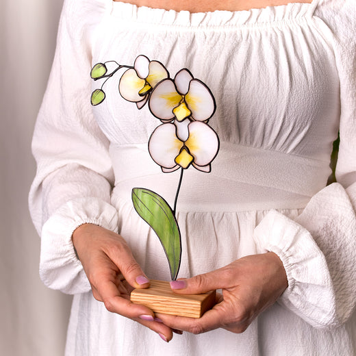 A person holding a stained glass artwork of a white orchid flower on a wooden platform.