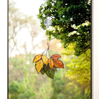 A handcrafted stained glass artwork featuring autumn leaves in shades of orange, yellow, and green, hanging in front of a window with a blurred background.