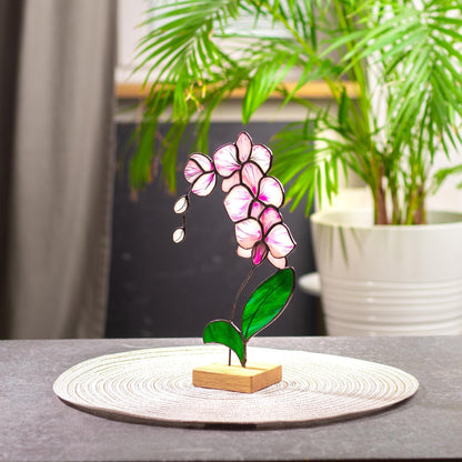 A handcrafted stained glass orchid with soft pink petals, delicate purple accents, and vibrant green leaves, mounted on a wooden platform.