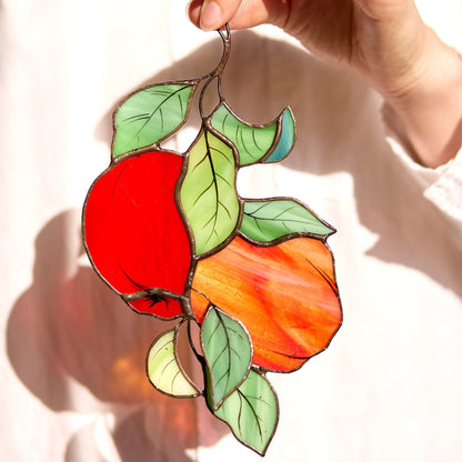 A handcrafted stained glass suncatcher in the shape of red apples with green leaves, designed to be hung by a window.
