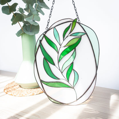 A stained glass suncatcher panel featuring a eucalyptus branch design with green leaves on a white background, hanging by a metal chain.
