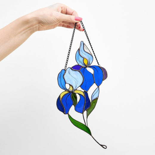 Hand holding a stained glass suncatcher in the shape of an iris flower with blue petals and green leaves.