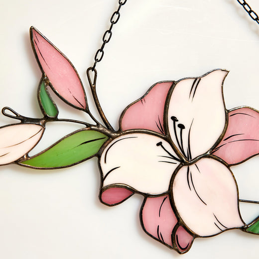 A handcrafted stained glass suncatcher in the shape of a pink and white lily flower with green leaves, soldered with copper foil and tin with black patina, hanging by a chain.
