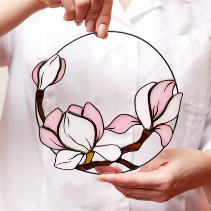 A person holding a round pink magnolia flower stained glass suncatcher.