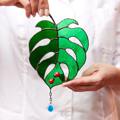 Hand holding a handmade stained glass suncatcher in the shape of a Monstera Deliciosa leaf with a red ladybug and a blue teardrop detail.