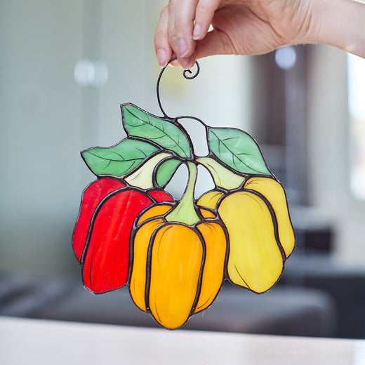 A handcrafted stained glass suncatcher in the shape of a pepper, with translucent red, yellow, and green panels, and a small hook at the top for hanging.
