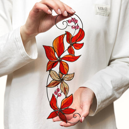 A hand holding a stained glass suncatcher with a design of red and orange parthenocissus leaves.