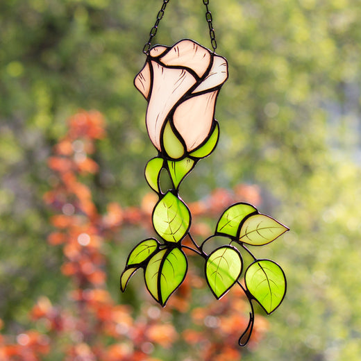 A handcrafted stained glass suncatcher in the shape of a rose flower with green leaves, hanging against a blurred background of autumn leaves.