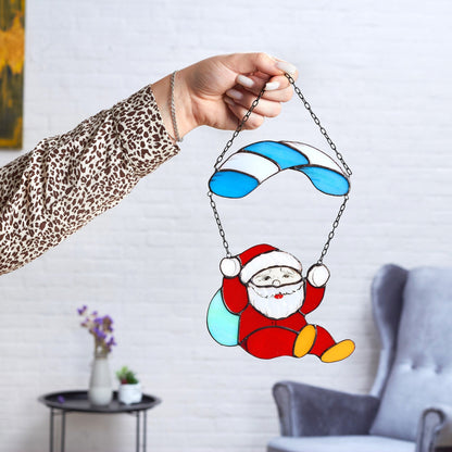 A hand holding a stained glass suncatcher depicting Santa Claus with a red suit and parachute in blue and white colors.