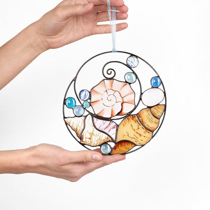 A handcrafted stained glass suncatcher featuring sea shell designs, held in hands against a light source to show the colorful patterns it casts.