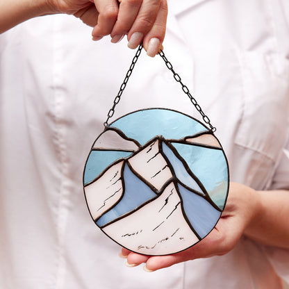 A person holding a round stained glass window hanging featuring mountain-like designs in blue and white colors with a chain attached for hanging.