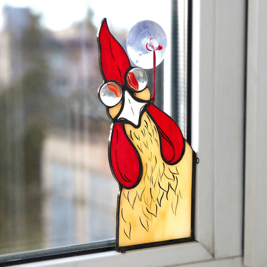 A stained glass window hanging of a red and yellow rooster with a whimsical expression, attached to a window with a vacuum suction cup.