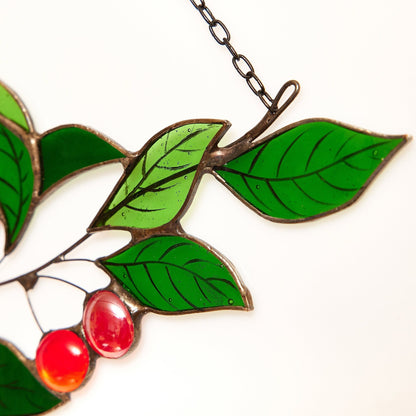 A handcrafted stained glass suncatcher featuring green leaves and red cherries, with a chain for hanging.
