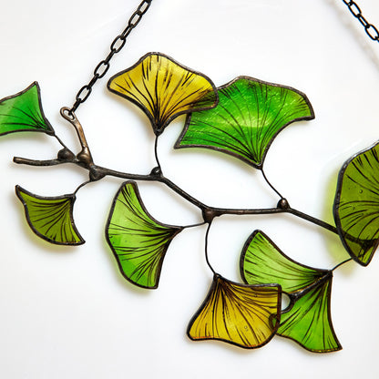 A handcrafted stained glass suncatcher in the shape of a Ginkgo Biloba leaf with green and yellow colors, soldered with black patina and hanging from a chain.