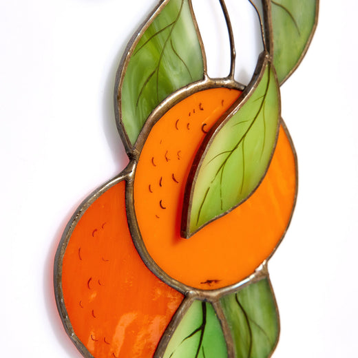 A handcrafted stained glass suncatcher in the shape of an orange tree branch with green leaves and orange fruits, with a black patina finish and copper foil.