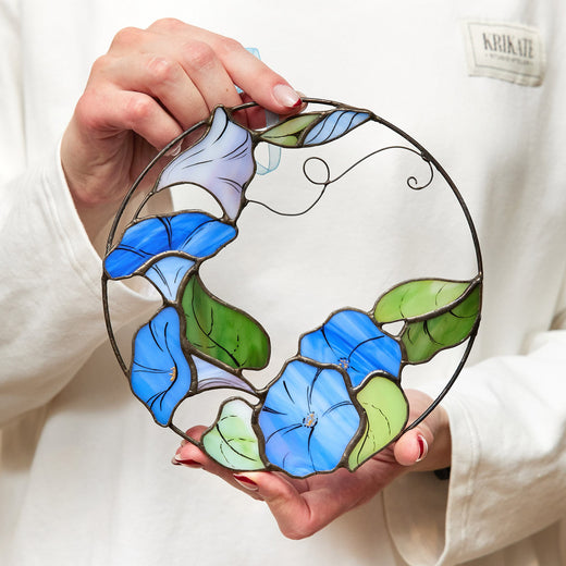 A hand holding a round stained glass panel with a morning glory flower design in blue and green colors.
